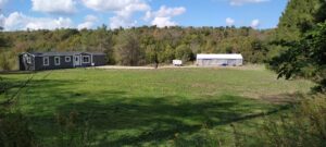 Wide view of rural property with a single-storey home, large outbuilding, and open green field surrounded by forest